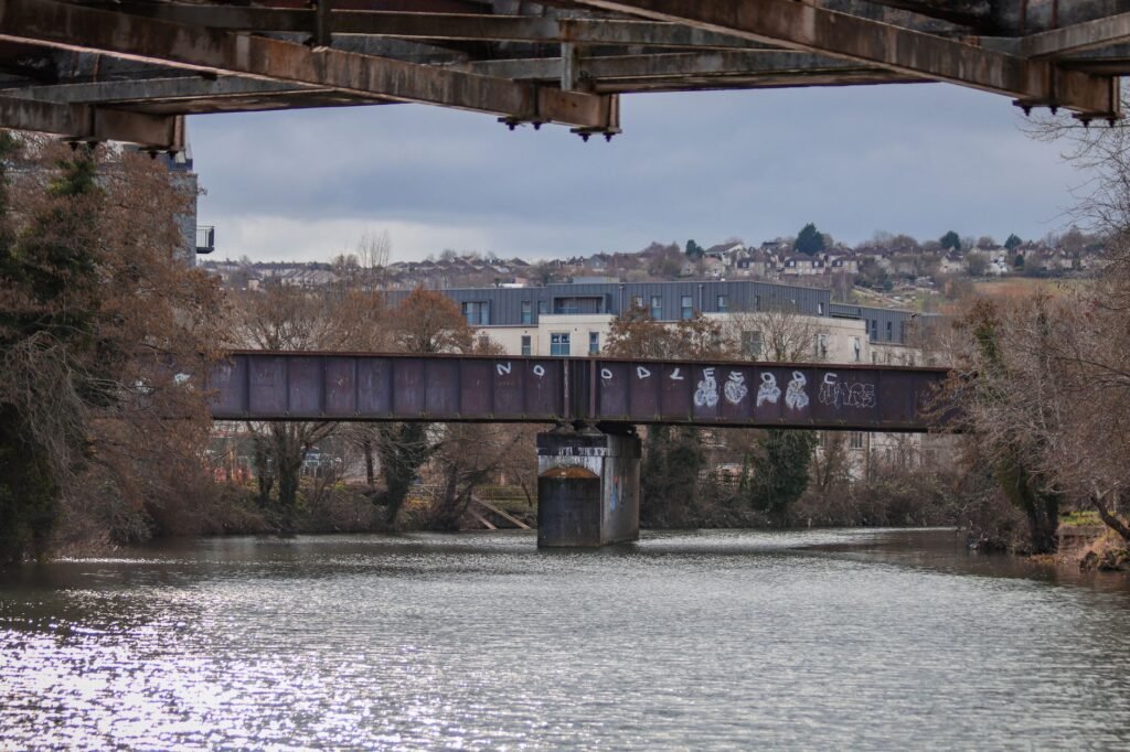A rusty metal bridge with graffiti spans over a tranquil river in Bath, England, under a cloudy sky.