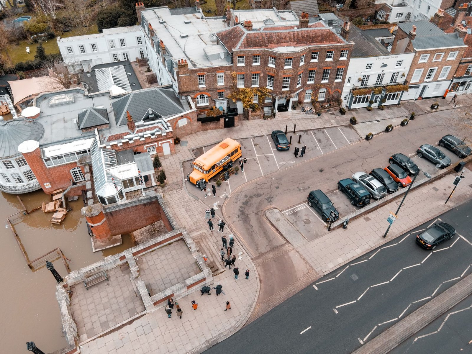 Drone view of school building facade near asphalt roadway and parking area with cars and bus with sidewalk and people near ri