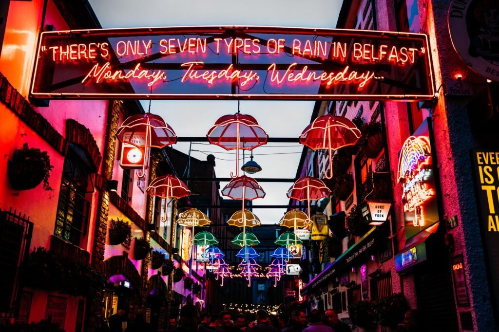 Bustling Belfast street illuminated by colorful neon umbrellas and a witty sign.
