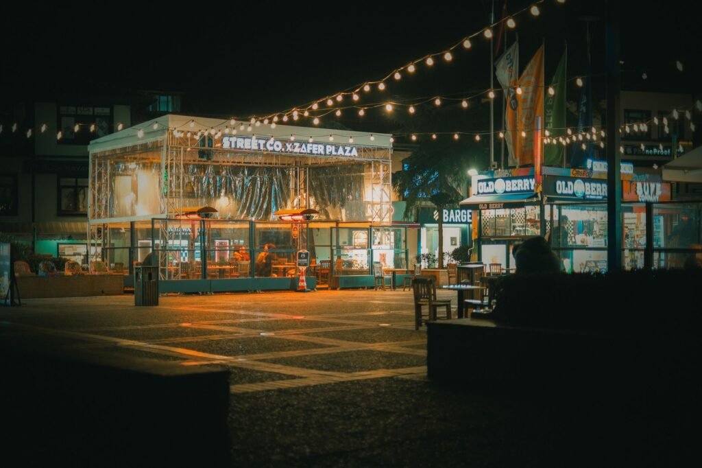 A bustling urban plaza illuminated by string lights at night, featuring lively shops and seating areas.