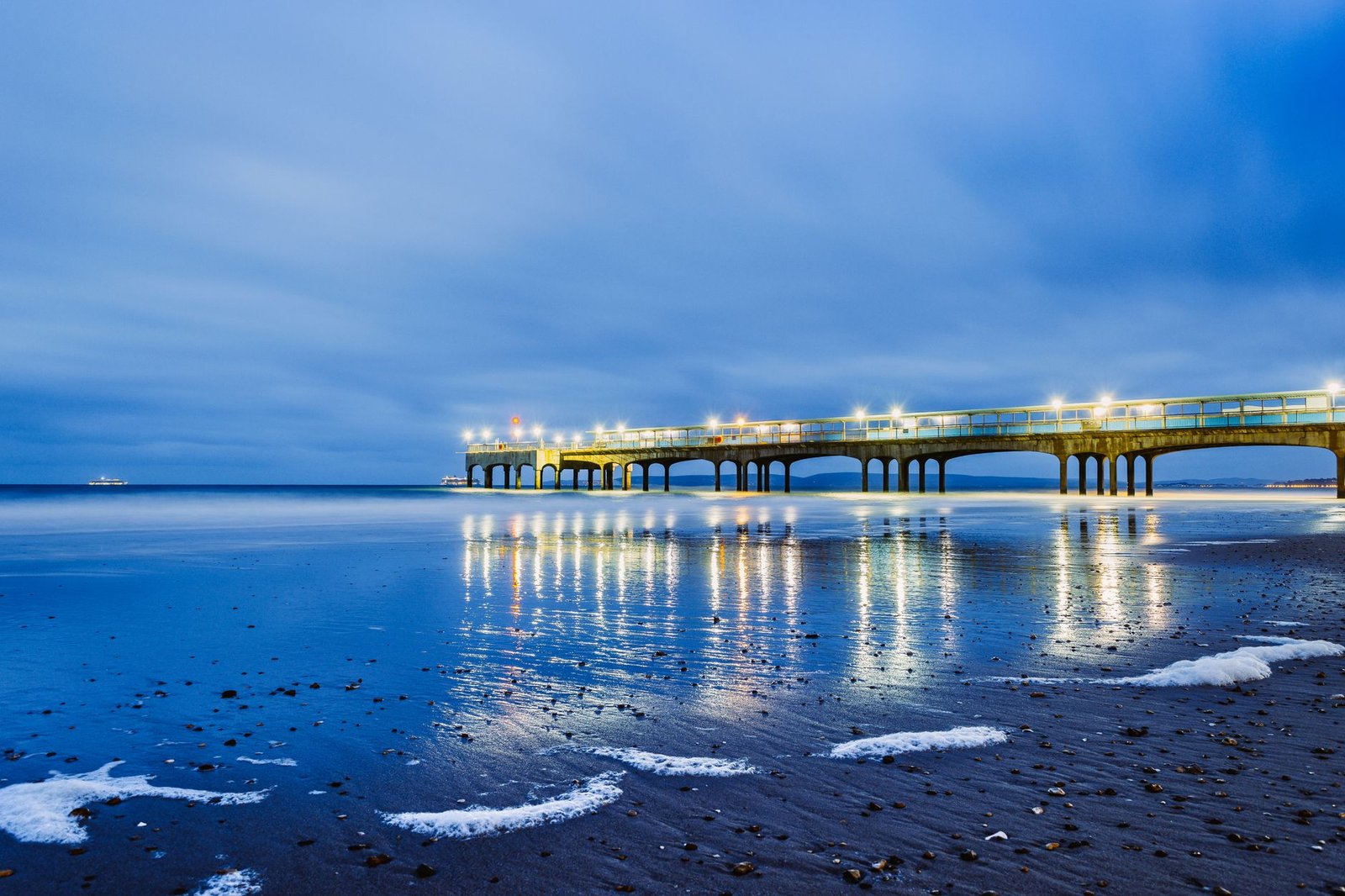 A serene view of the illuminated Boscombe Pier reflecting on calm waters during dusk in Bournemouth, England.
