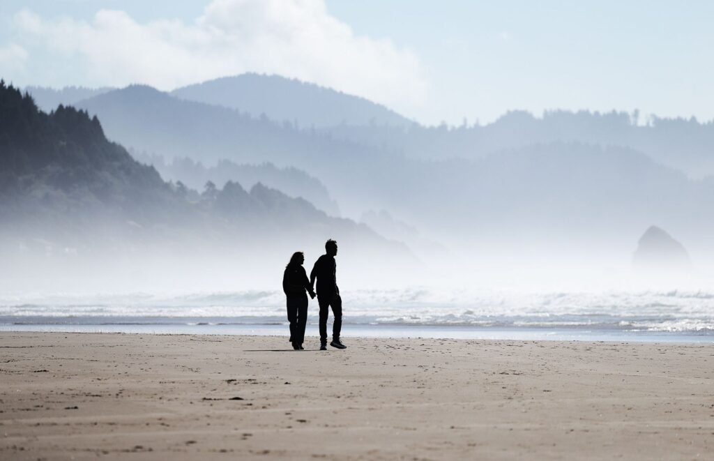 Romantic couple holding hands walking along Cannon Beach with coastal backdrop.