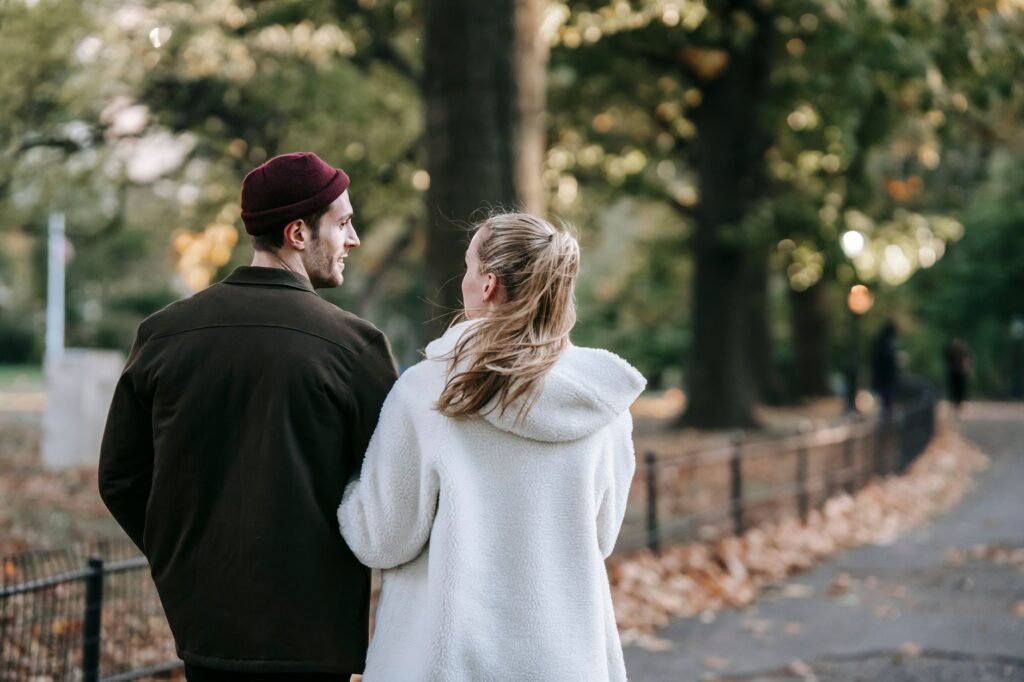 Back view of young romantic couple in stylish outfits holding hands and communicating while walking on alley in autumn park o