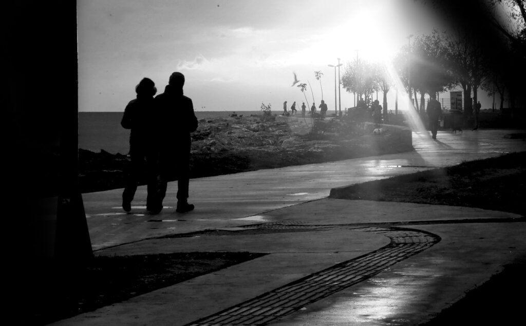 Black and white photo of a couple walking on a sunlit promenade by the sea.