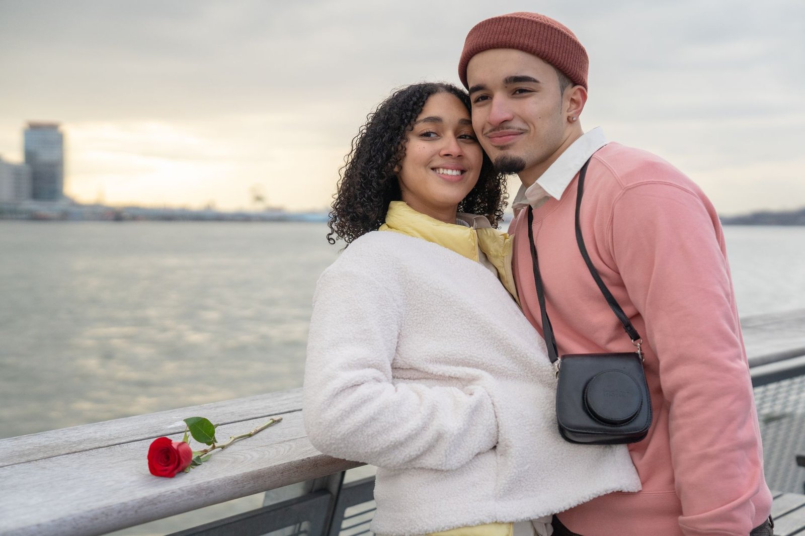 Cheerful Hispanic couple with photo camera looking away while standing near railing on waterfront near sea during romantic da