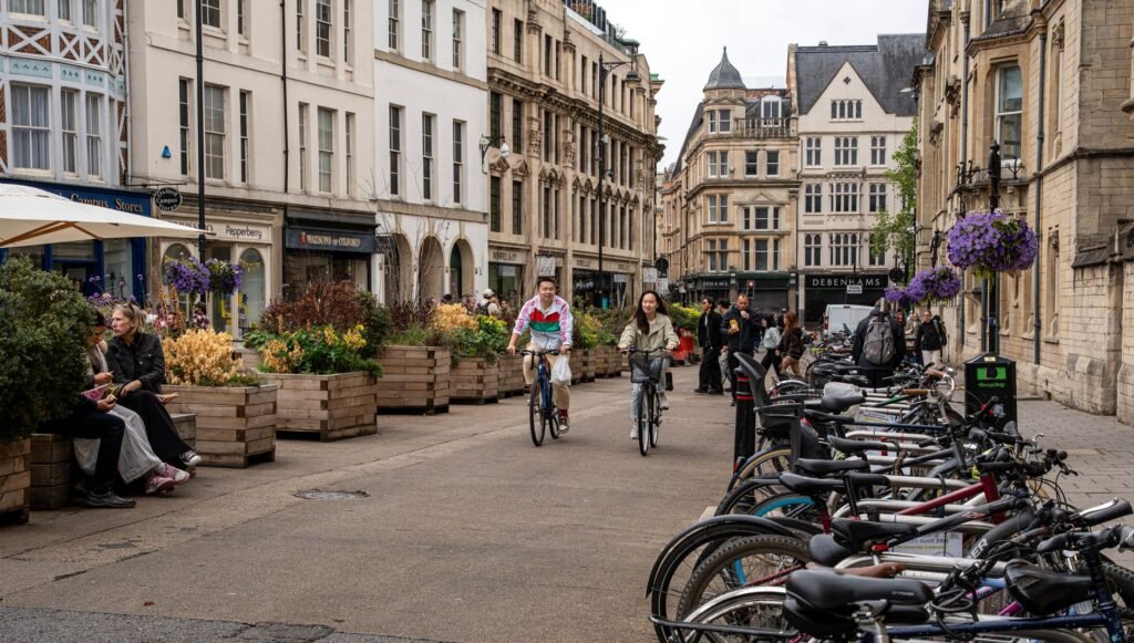 People bicycling and enjoying a lively street in Oxford with historic architecture.