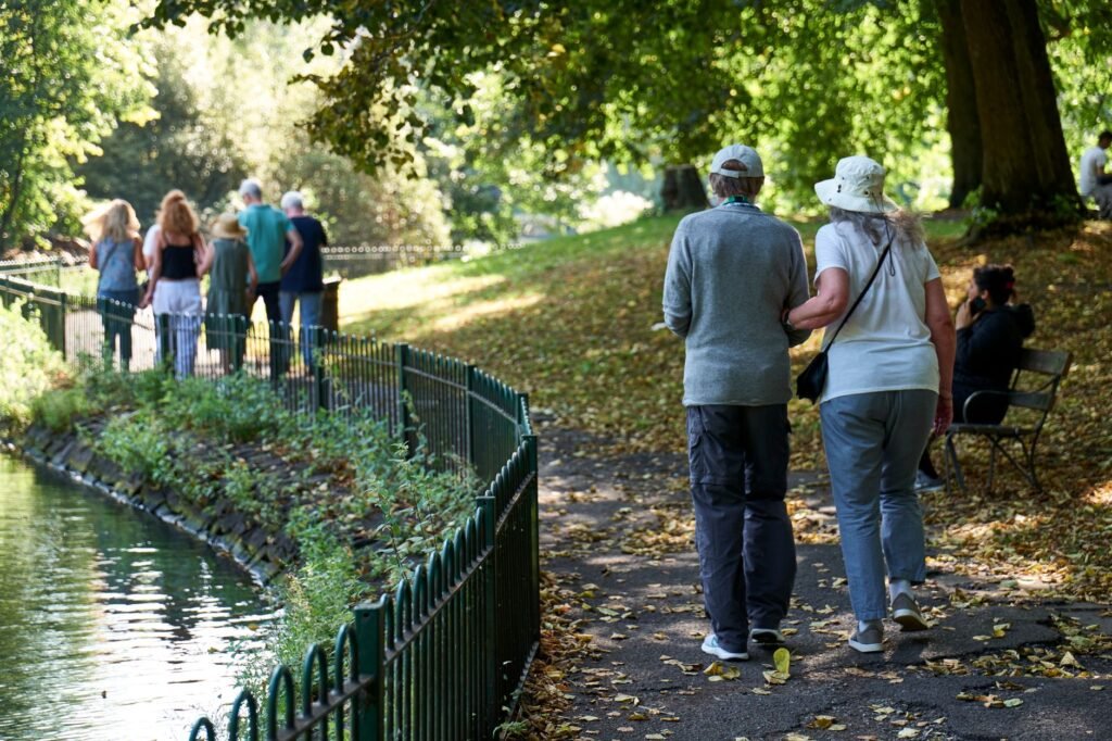 Back view of unrecognizable elderly couple in casual outfits holding hands and strolling along pond in park in sunlight