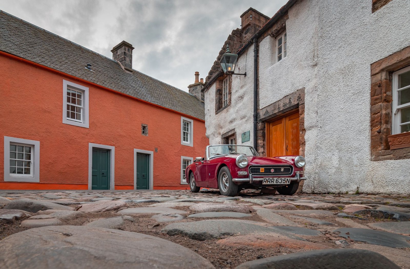 A classic vintage red convertible parked on cobblestone in Culross, Scotland, amidst historic buildings.