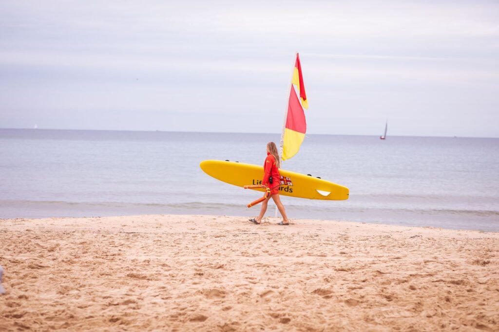 A lifeguard in red uniform walking with a yellow surfboard on Bournemouth beach.