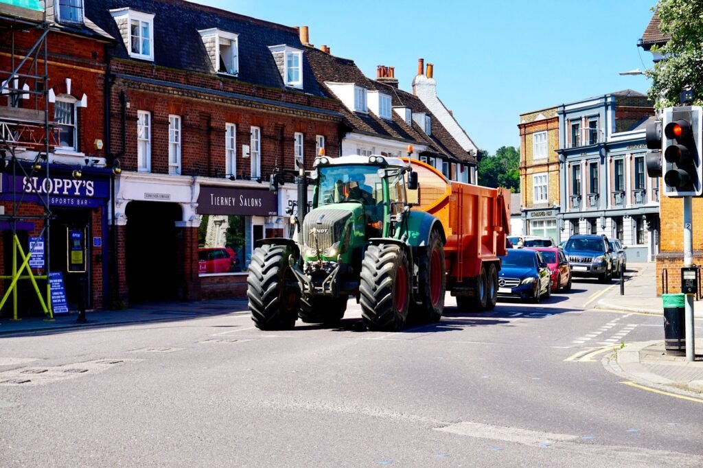 A green tractor drives through Hertford town center, surrounded by cars and shops.
