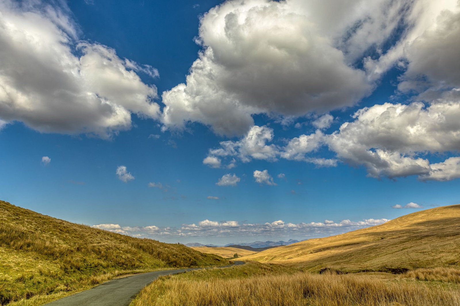 A picturesque Scottish countryside with a winding road under a vibrant blue sky and fluffy clouds.