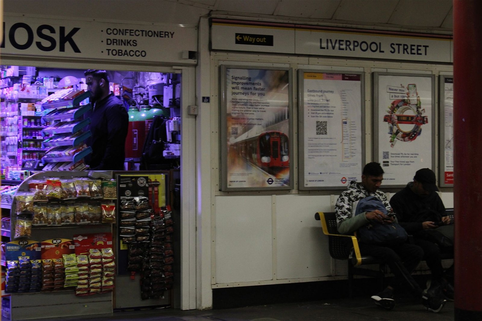 Night scene at Liverpool Street Station with a vibrant kiosk and waiting passengers.