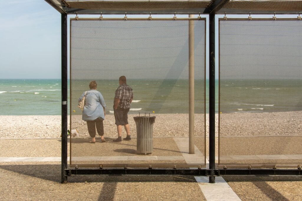 A couple enjoys a beach view through a metallic mesh barrier under a clear sky.