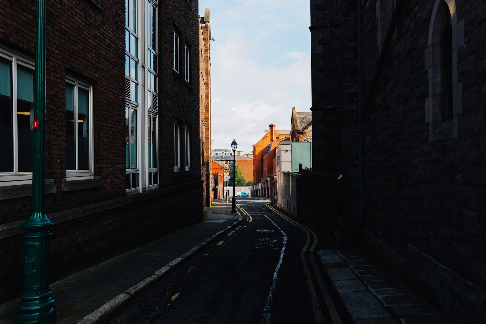 A narrow alleyway flanked by rustic brick buildings, leading to a sunlit street beyond.
