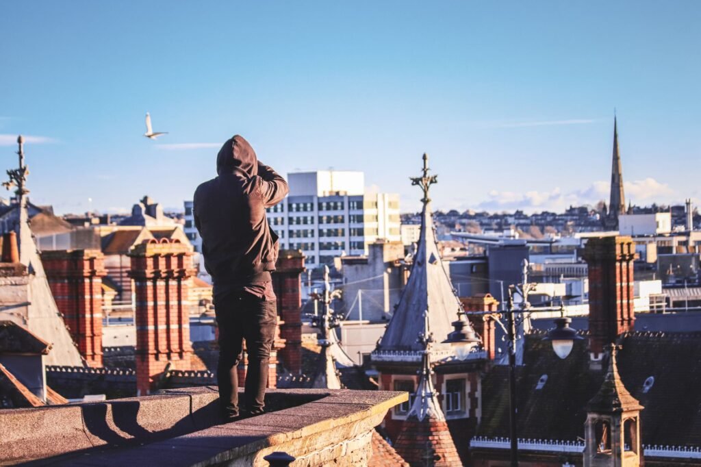 A man in a hoodie stands on a rooftop, capturing the scenic view of Bristol, England with city chimneys and spires in the bac
