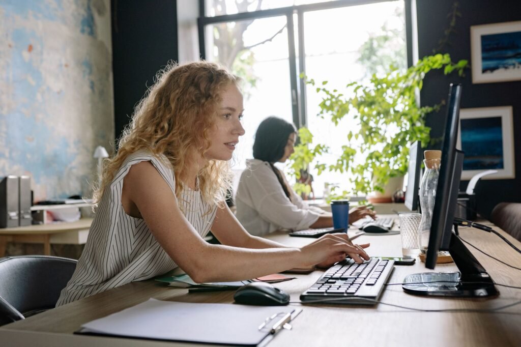 Two focused women working at a bright and modern office space, typing on computers at a shared desk.