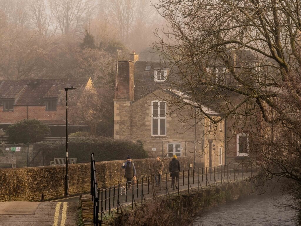Three people and a dog walking by historic stone houses on a foggy morning by the riverside.