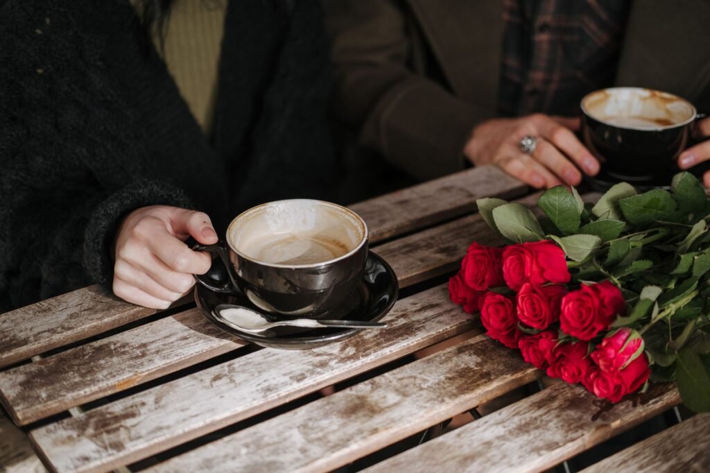 From above of crop anonymous couple with cups of coffee resting at wooden table with red roses in cafeteria