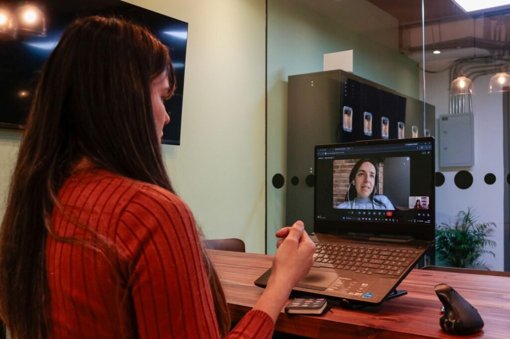Two women in a video call meeting, one in an office and the other on a laptop screen.