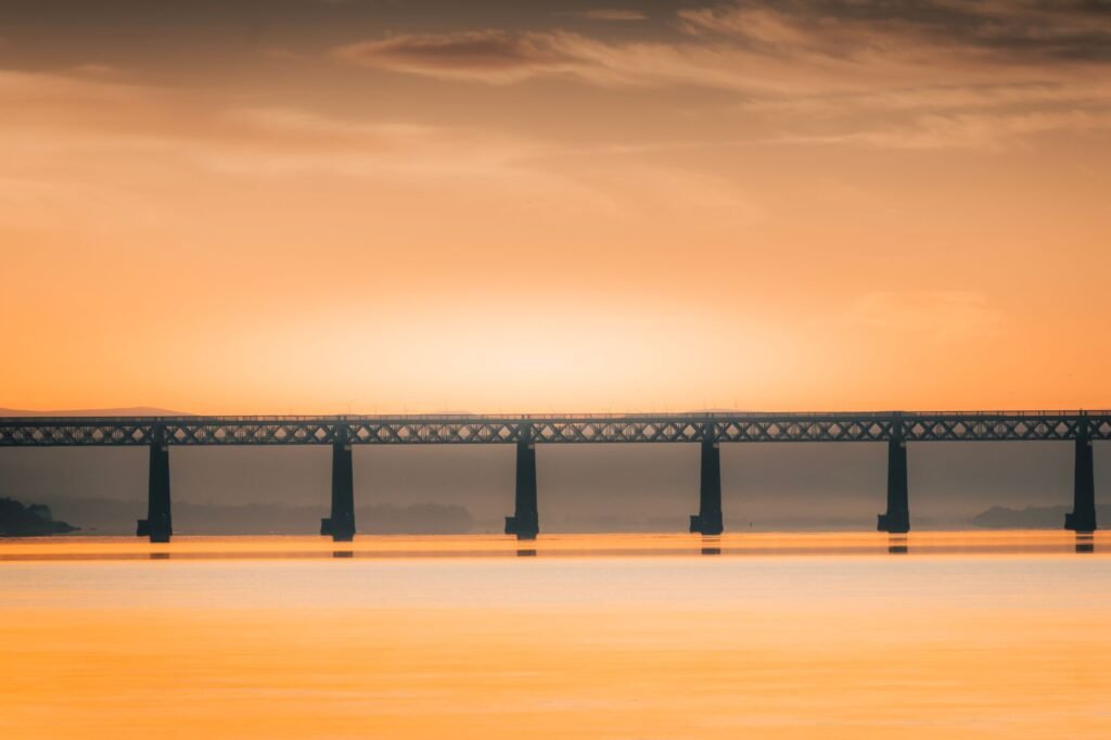 A vibrant sunset casts an orange glow over a railway bridge in Scotland, creating a serene landscape.