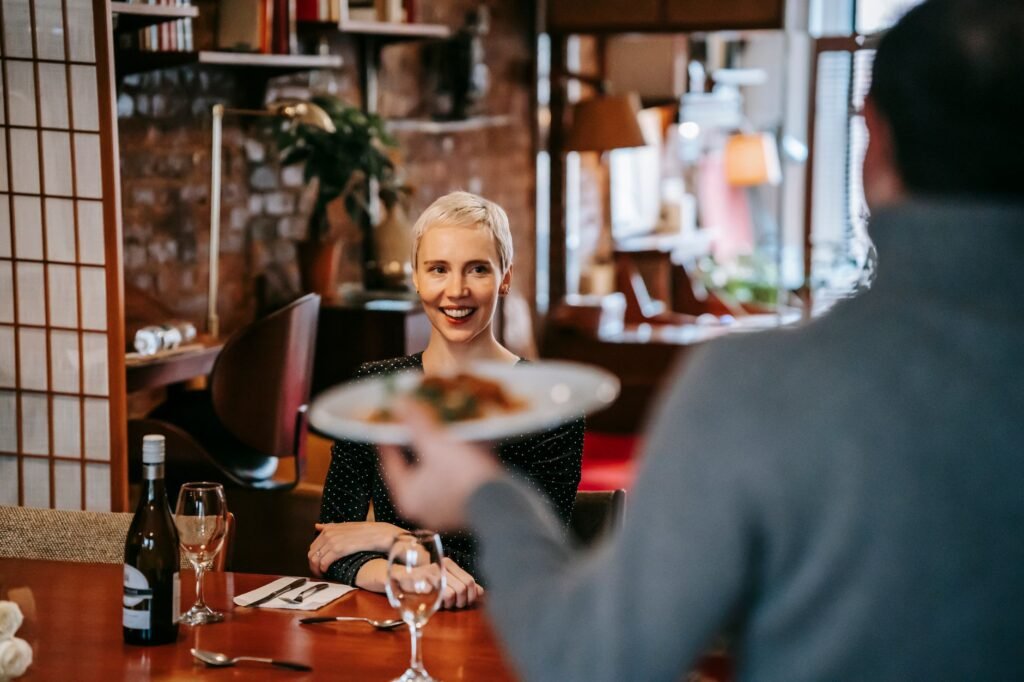 Positive couple having date in bright restaurant while man serving plate with food to woman sitting in chair at table with wi
