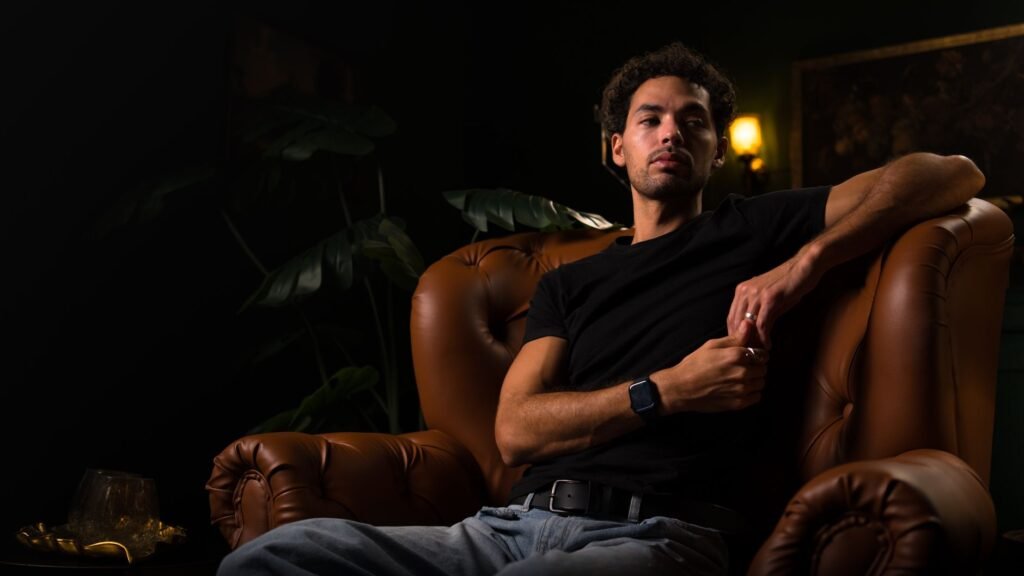 A fashionable man in casual attire relaxes on a leather chair in a dark and stylish setting.