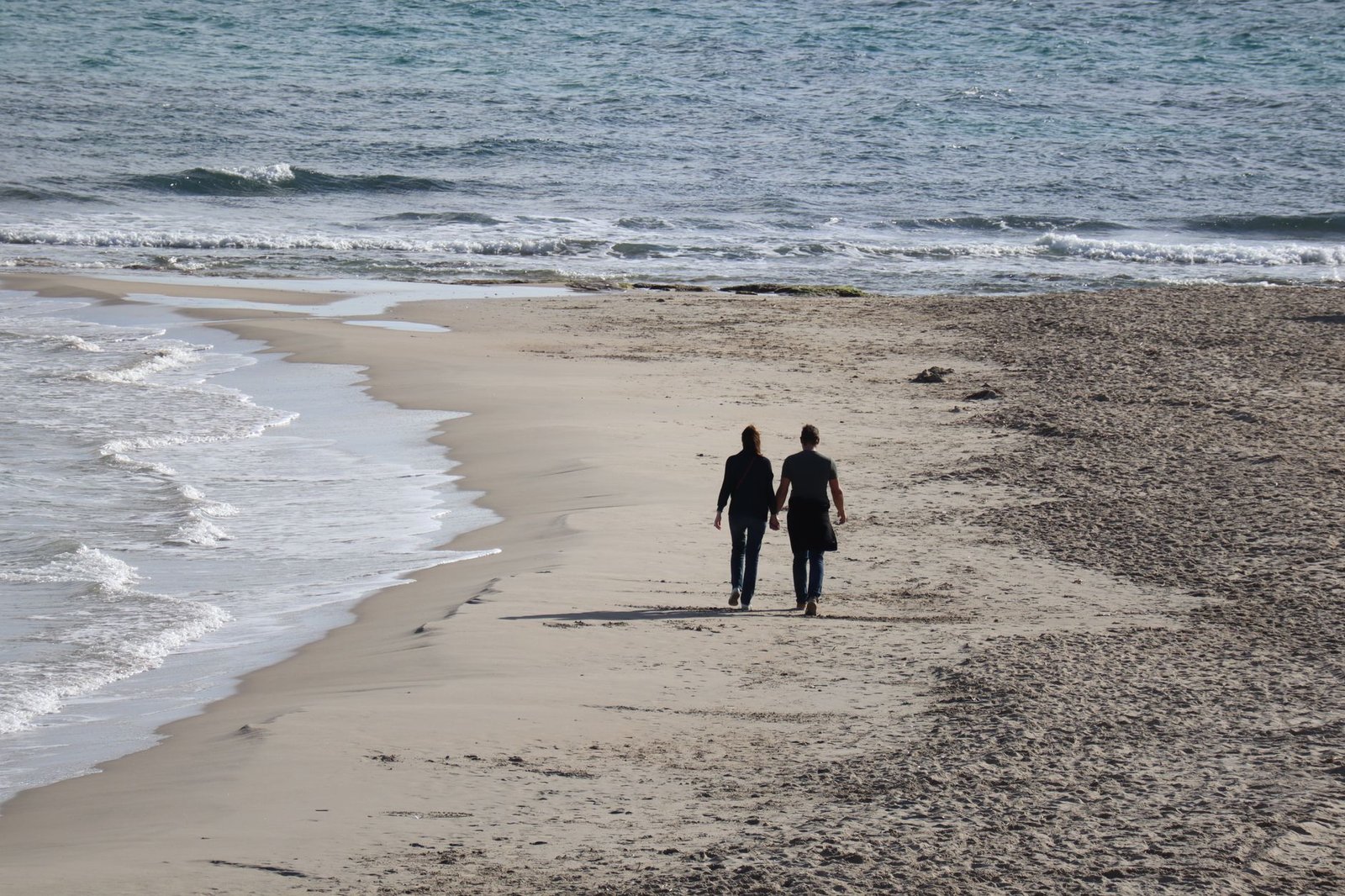 A couple walking hand in hand on the sandy beach of Alicante, offering a serene seaside view.
