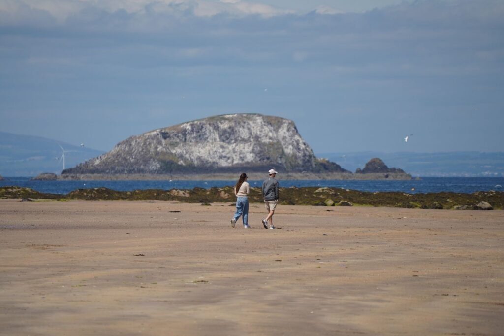 A couple strolls along a sandy beach with Bass Rock in the background, North Berwick, Scotland.