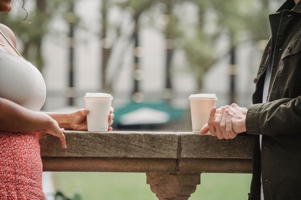 Side view of crop anonymous young couple in casual clothes standing on balcony with cups of coffee to go and chatting