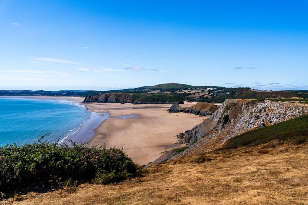 Aerial view of Rhossili Bay's expansive sandy beach and cliffs under a clear blue sky.
