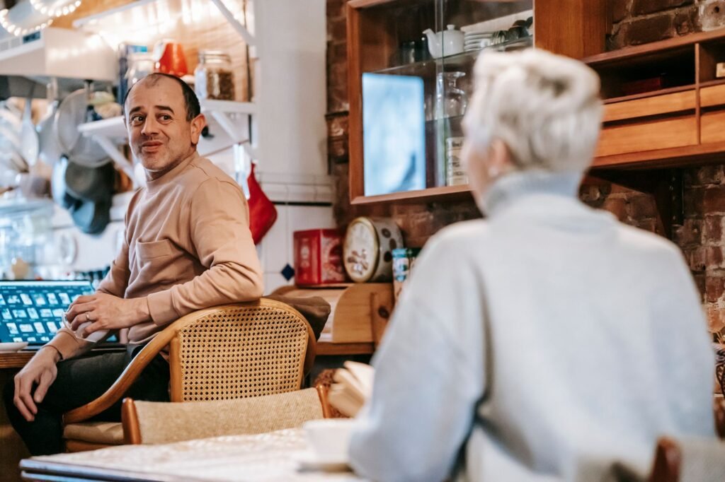 Side view of happy adult multiracial couple in casual clothes drinking coffee and chatting sitting at tables in apartment dur