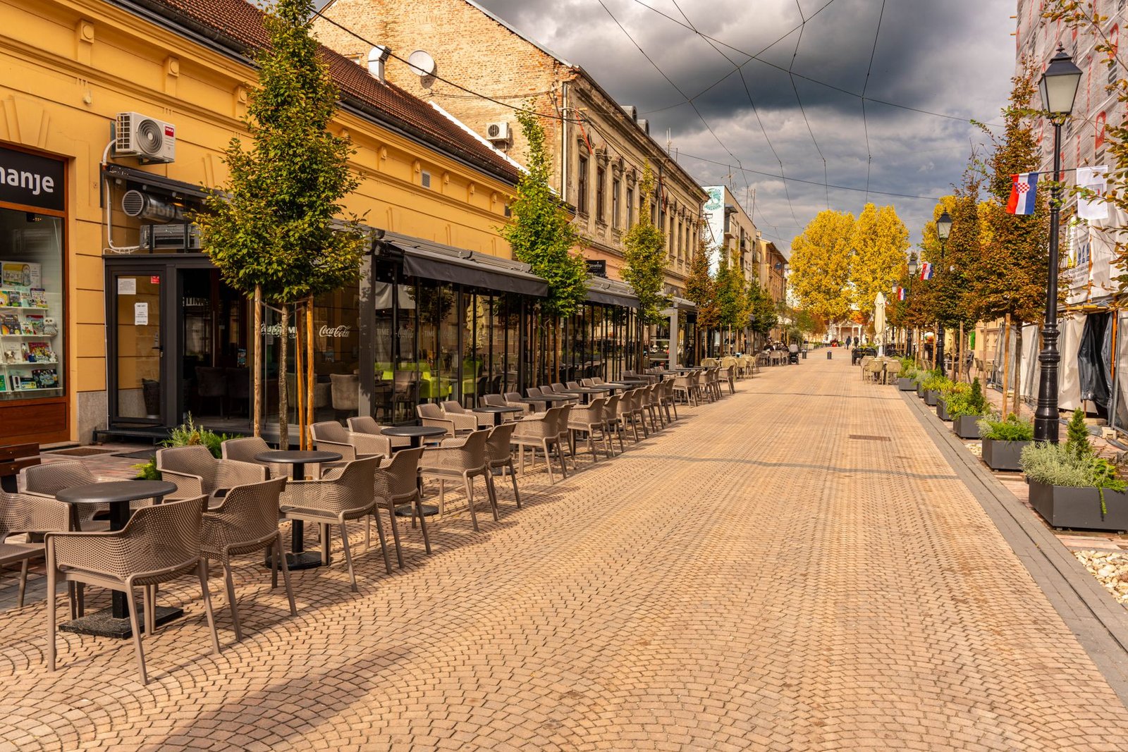 Empty European street with cafes and autumn trees under cloudy skies.
