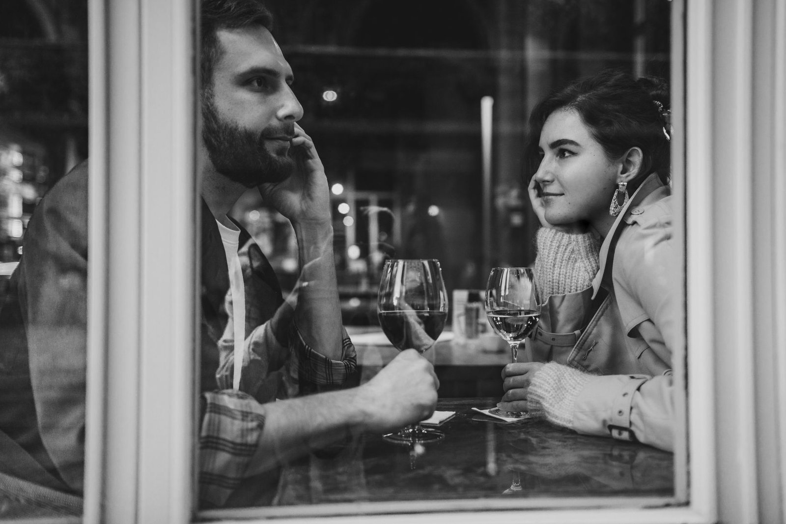 A couple enjoys a romantic evening with wine at a cozy cafe, captured through the window.