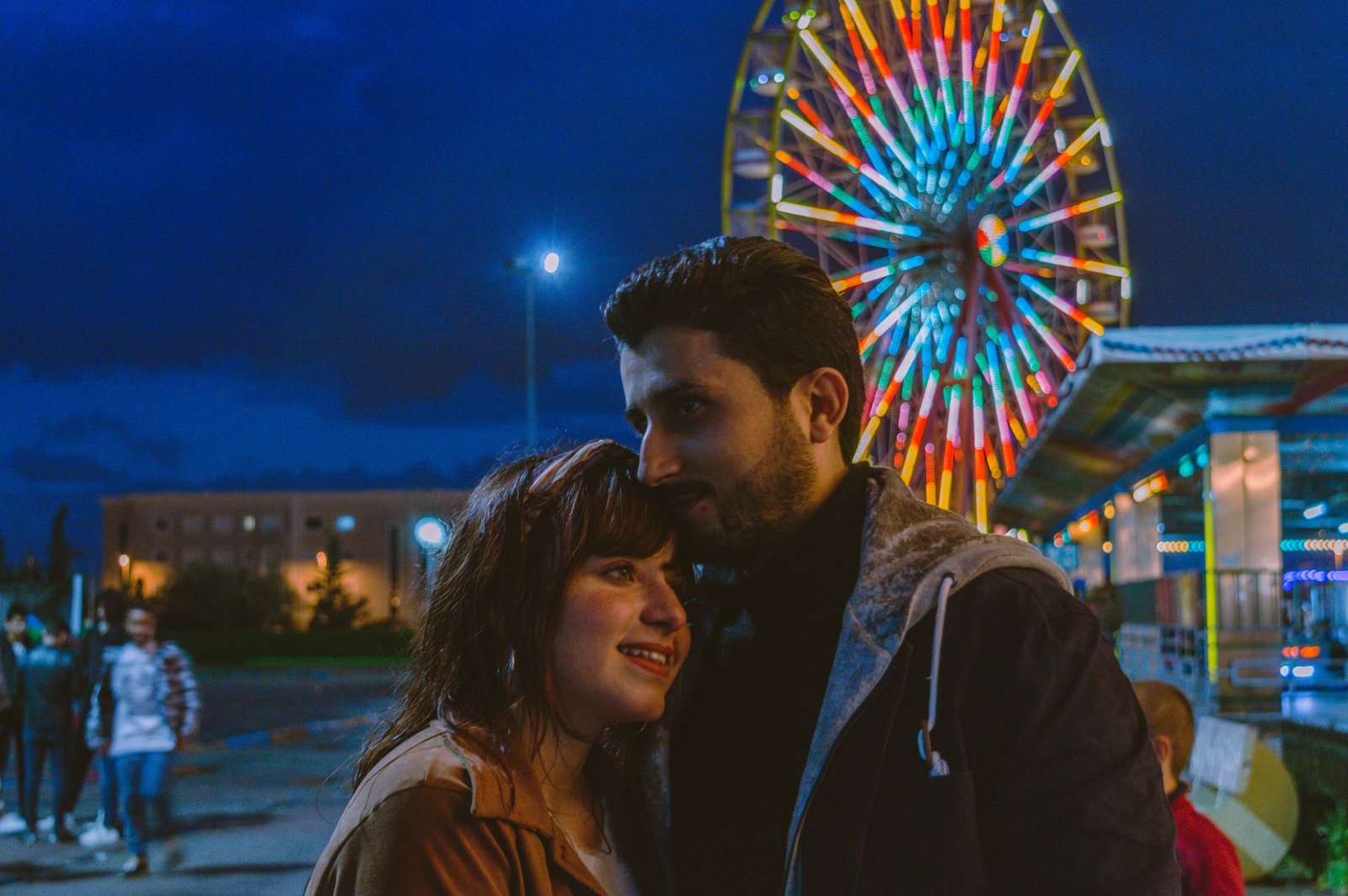 A couple shares a loving moment in front of a lit Ferris wheel at an amusement park.