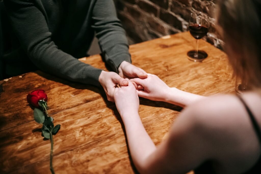 High angle of unrecognizable couple sitting at table in cafe with rose flower and glass of red wine and holding hands