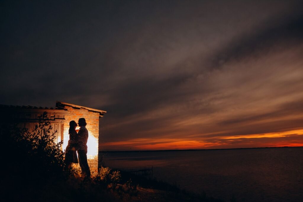 Silhouette of a couple embracing by a seaside shed at sunset, under a dramatic cloudy sky.
