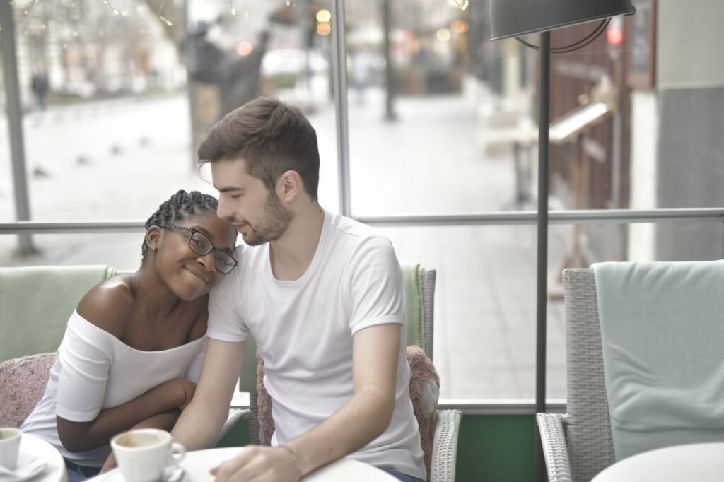 Content African American young woman in casual wear putting head to boyfriends shoulder and looking at camera while sitting i