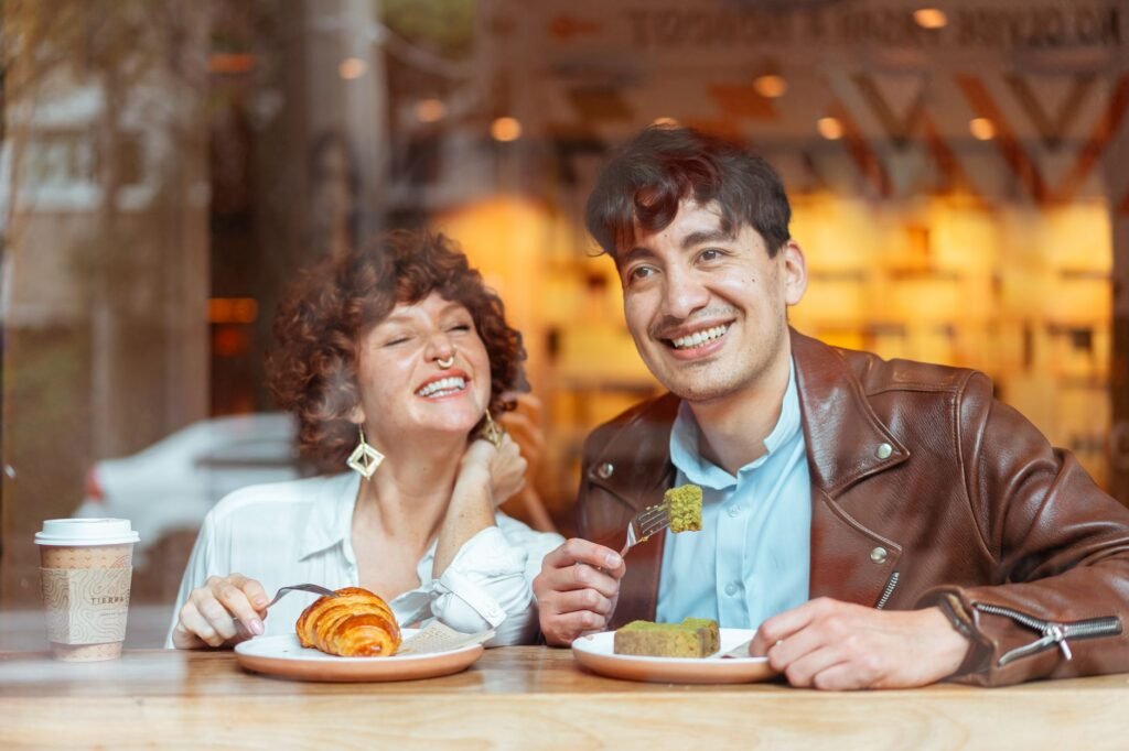 Interracial couple laughing and eating pastries at a cozy cafe.