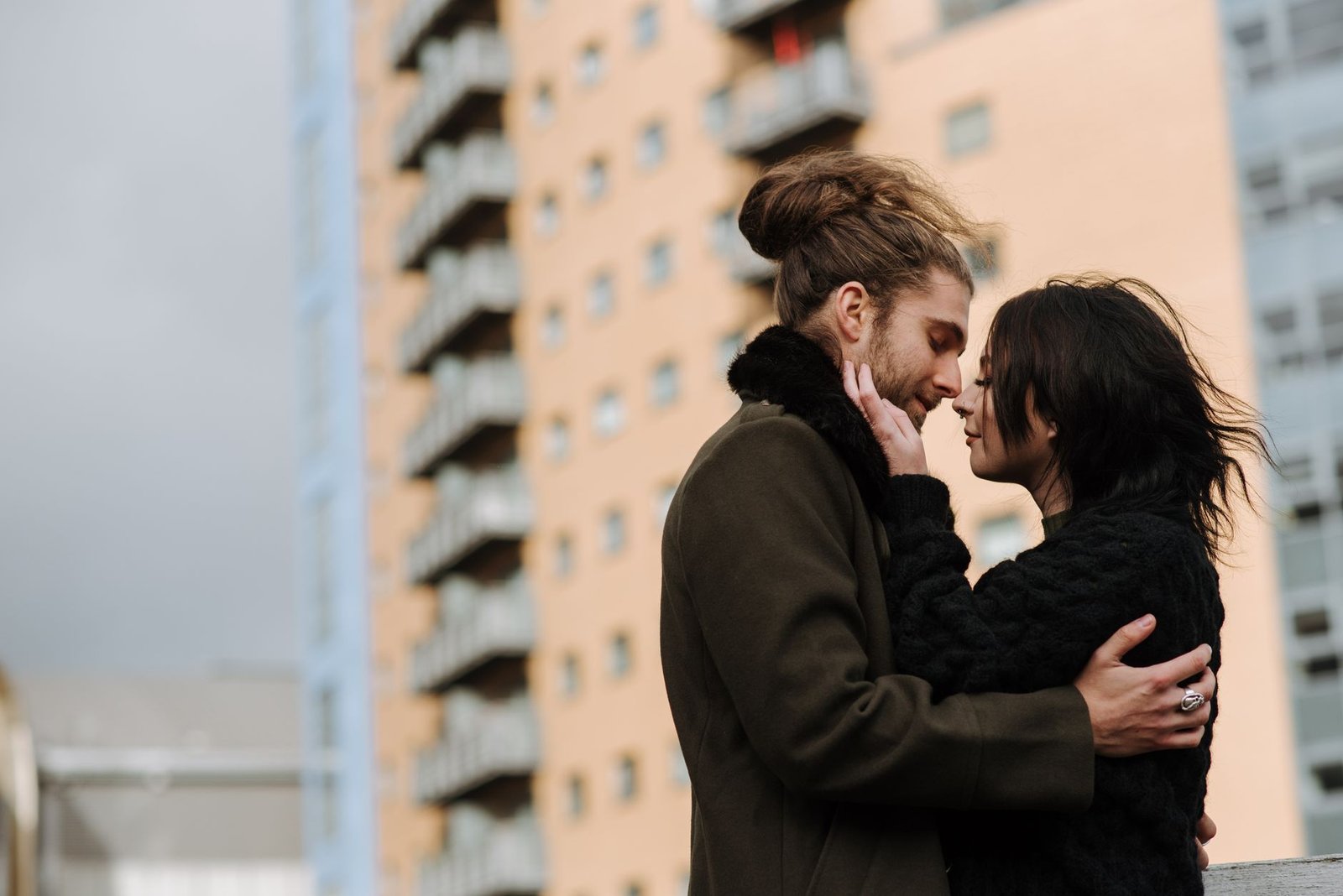 From below side view of romantic boyfriend and girlfriend hugging while standing on street in windy weather against building