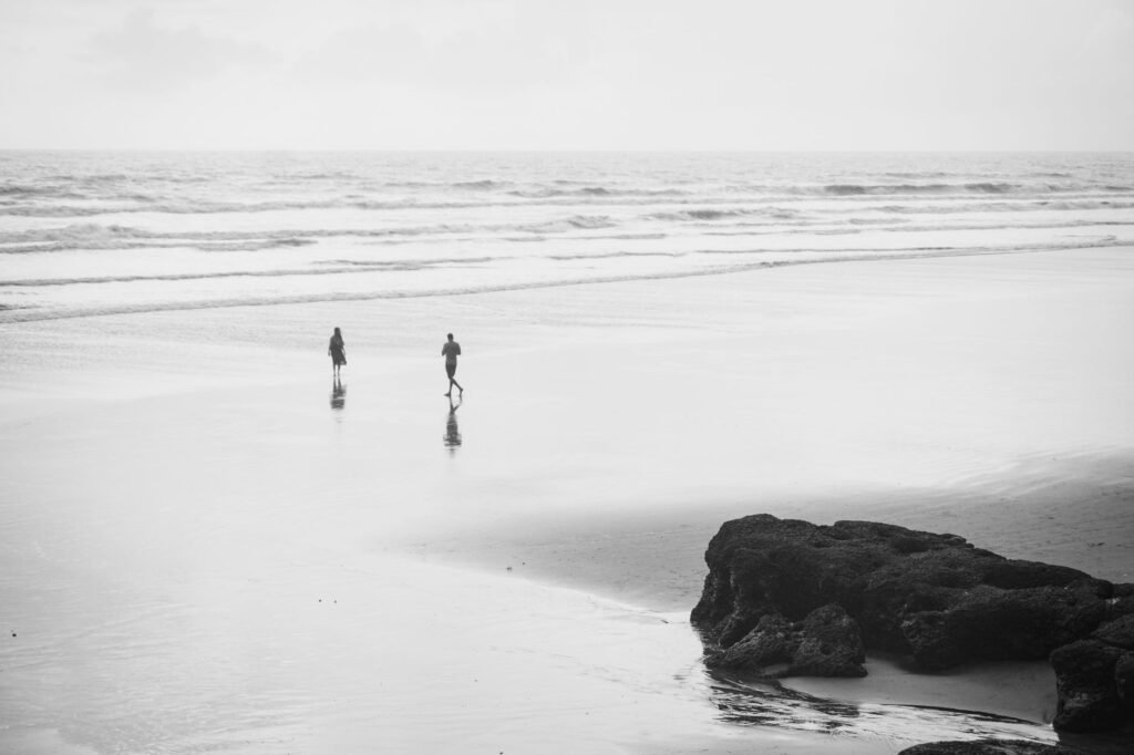 Two people walking on a serene beach in Morjim, India, captured in black and white.