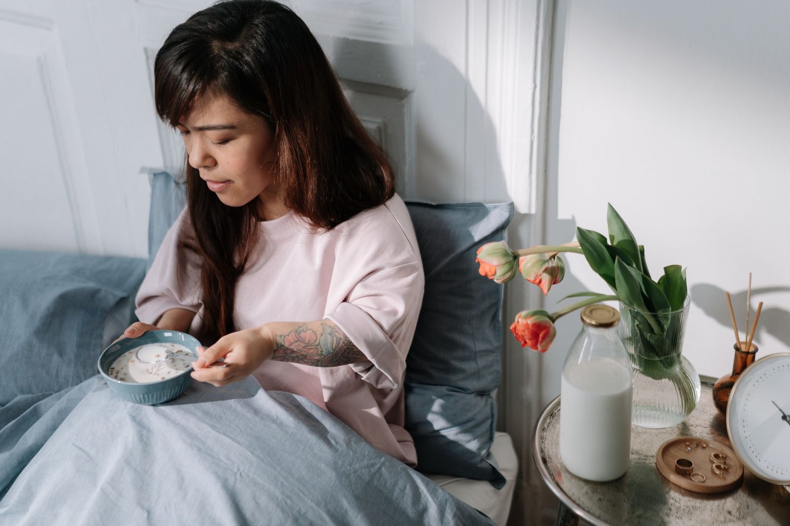 A woman sits in bed enjoying breakfast with milk and flowers nearby, bathed in morning light.