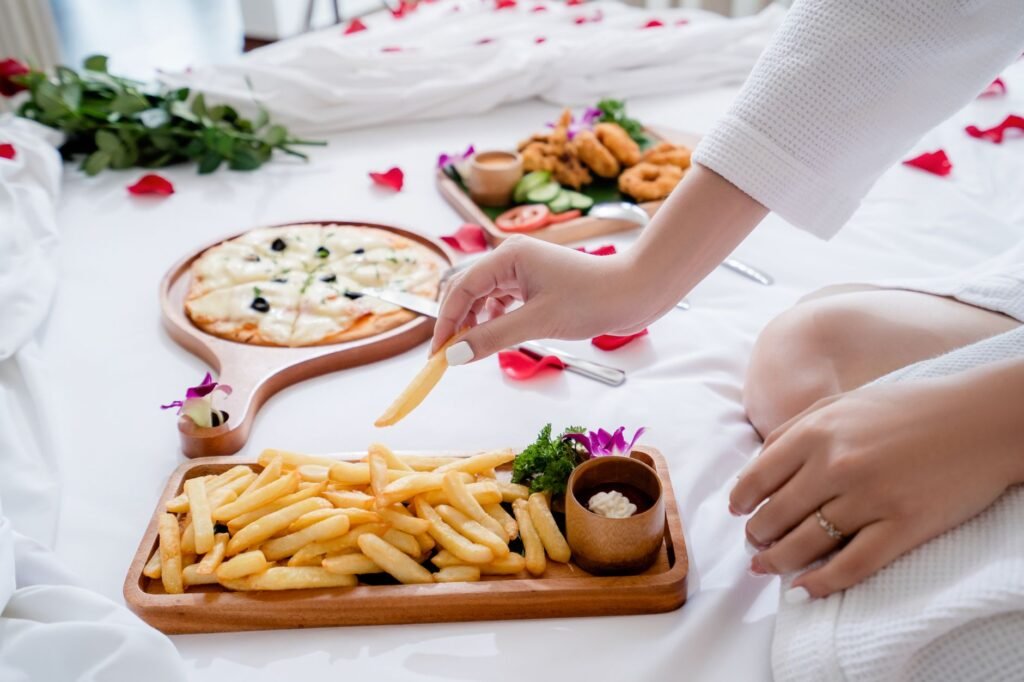 Enjoy a cozy breakfast in bed featuring crispy fries and gourmet pizza on a white sheet adorned with rose petals.
