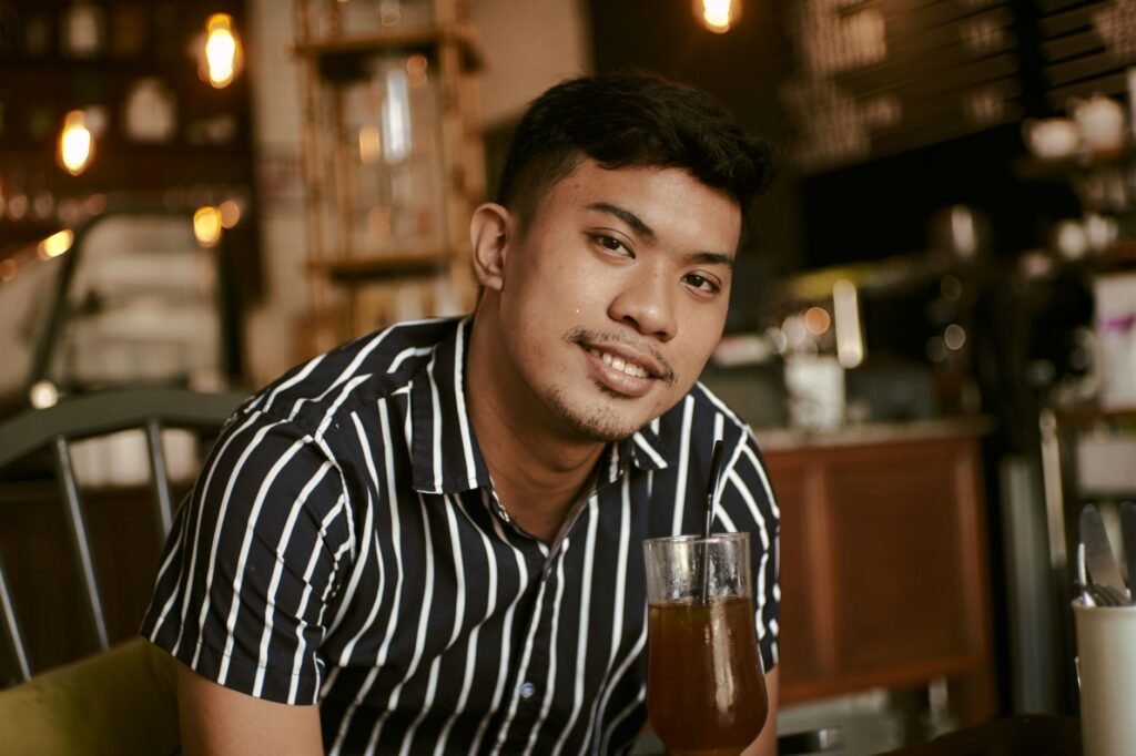 A smiling Asian man in a striped shirt enjoys a drink in a cozy coffee shop.