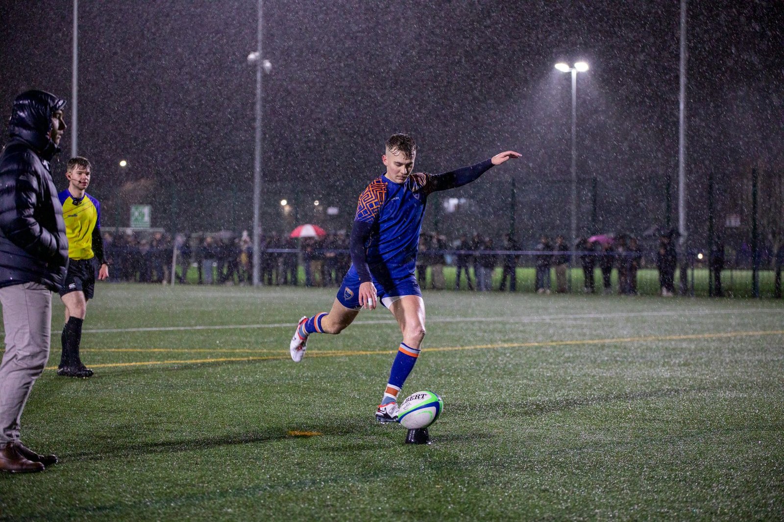 Dynamic shot of a rugby player kicking the ball during a night match in the rain at a Manchester stadium.