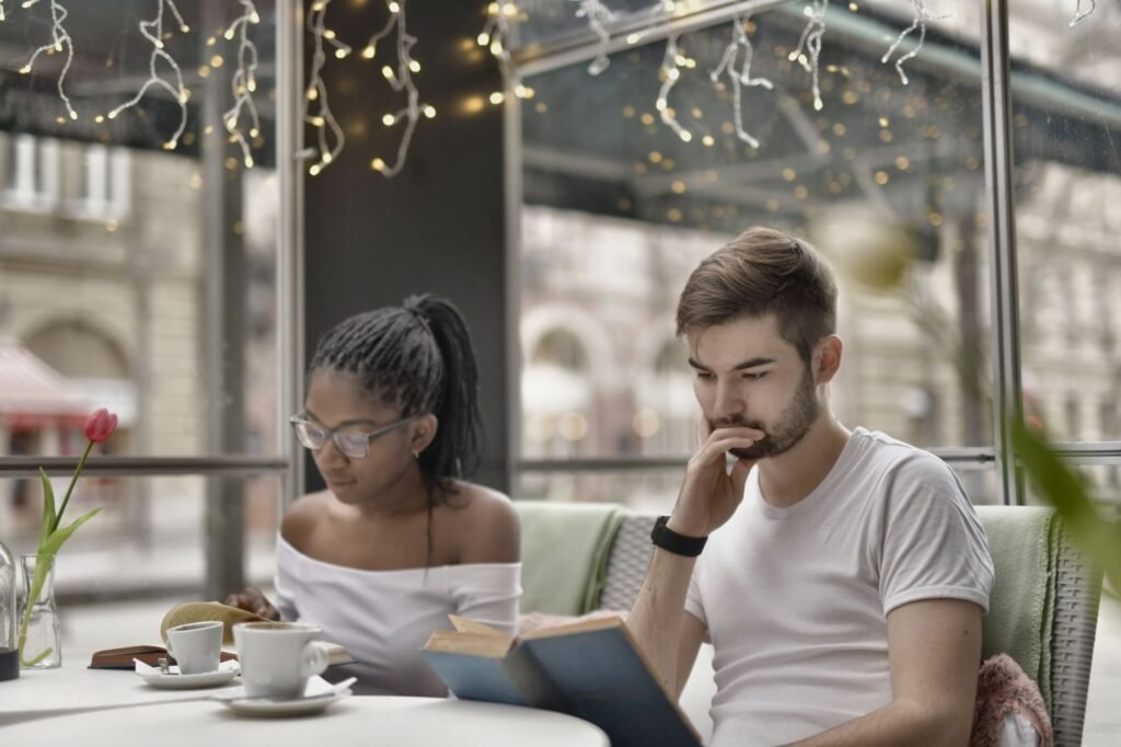Pensive young African American woman in eyewear sitting near wistful friend in smart watch while reading books together and d