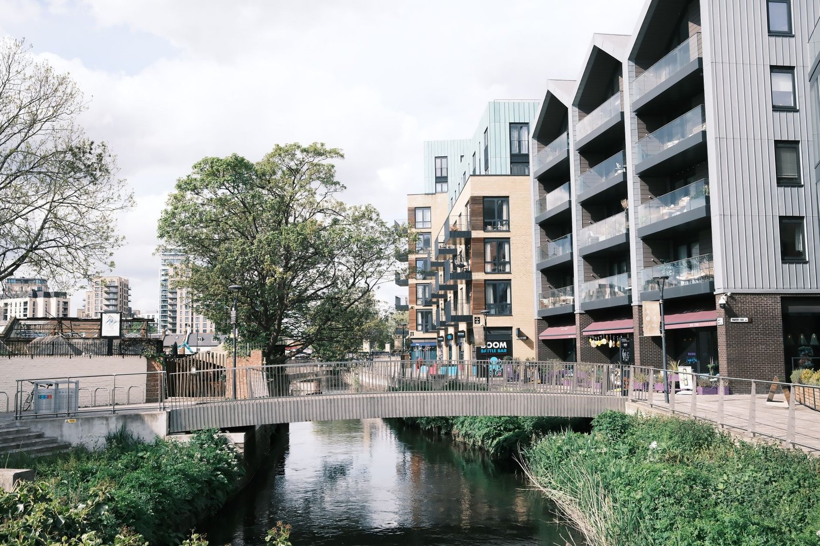 Contemporary apartment buildings line a calm river with a pedestrian bridge in the foreground.