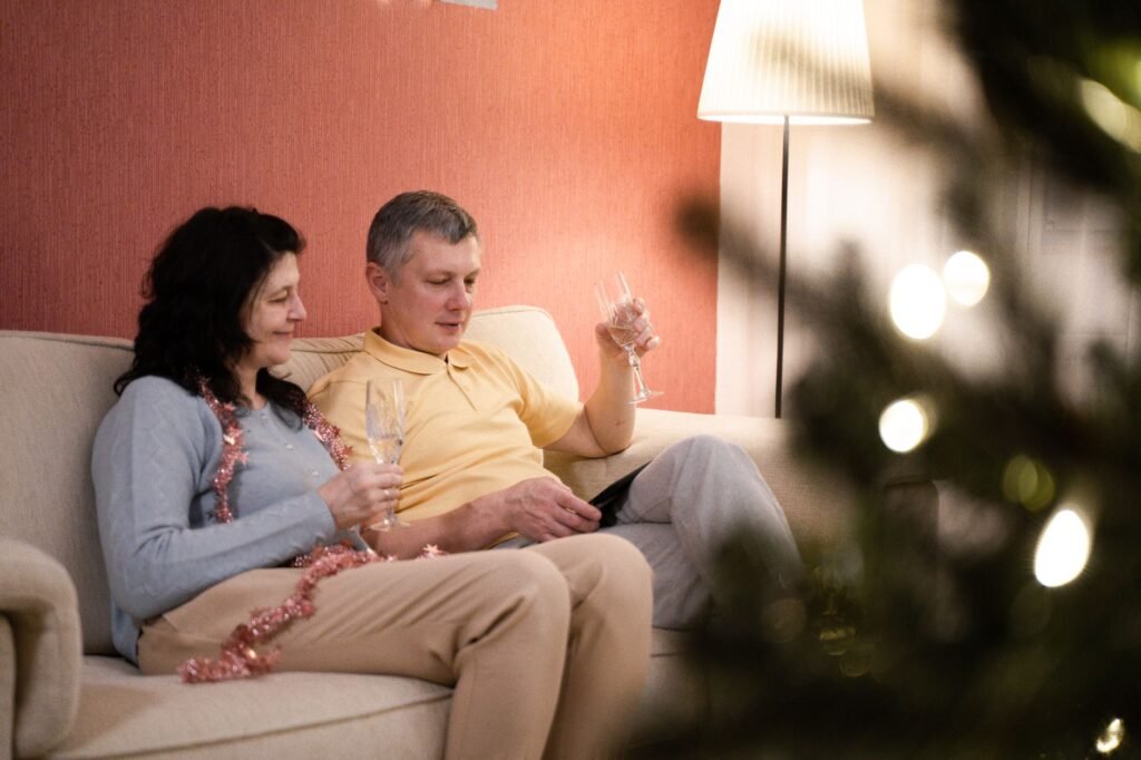 A couple enjoying a cozy evening, toasting with champagne on a couch with a festive tree nearby.