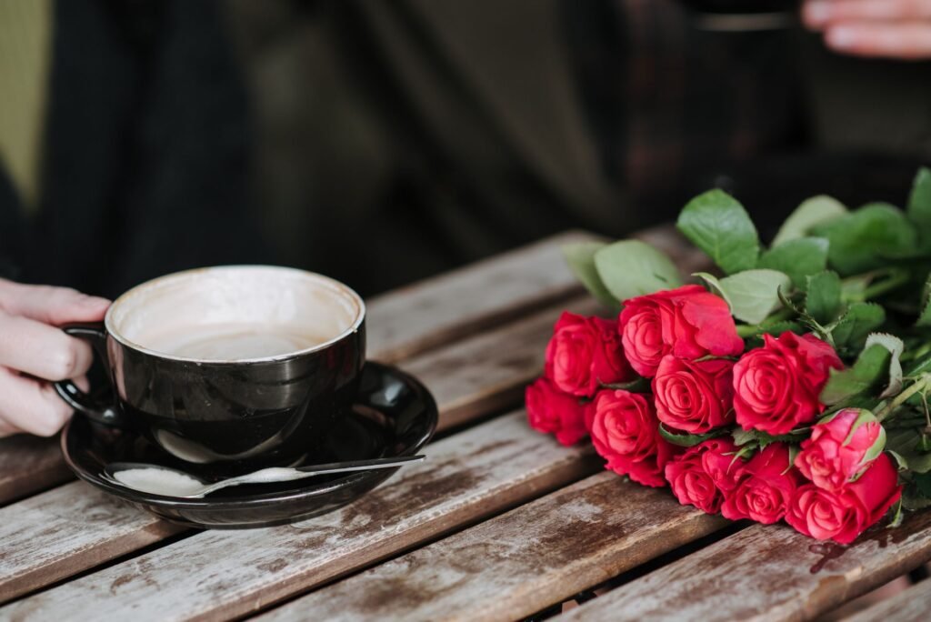 Crop unrecognizable couple with cup of delicious cappuccino at wooden table with blooming red flowers