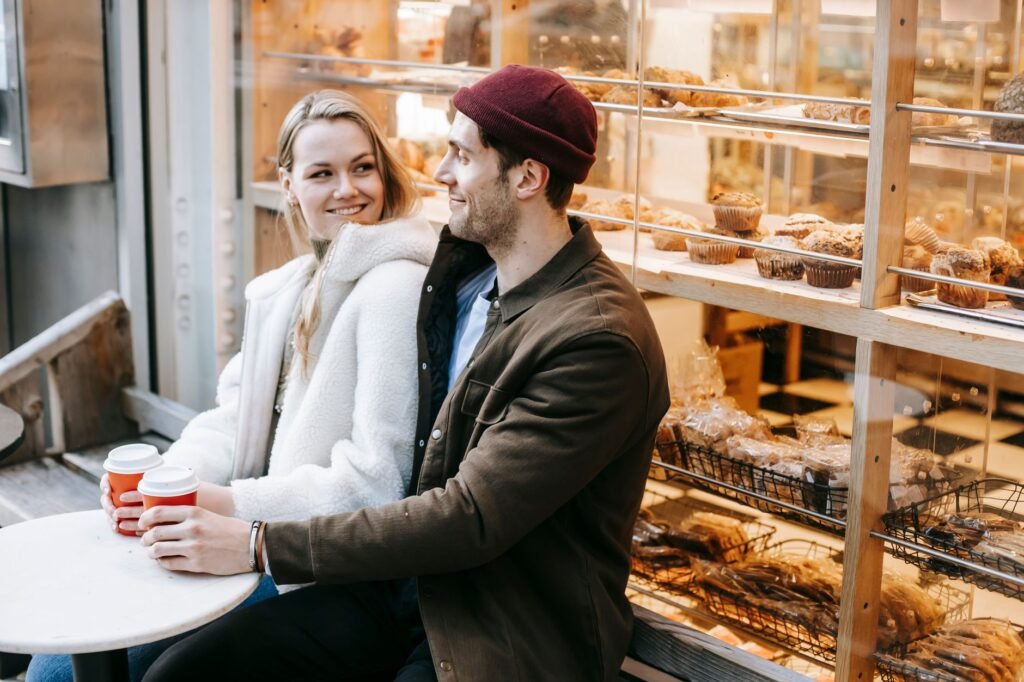 Cheerful young couple in casual clothes sitting at table with paper cups with hot beverage and looking at each other