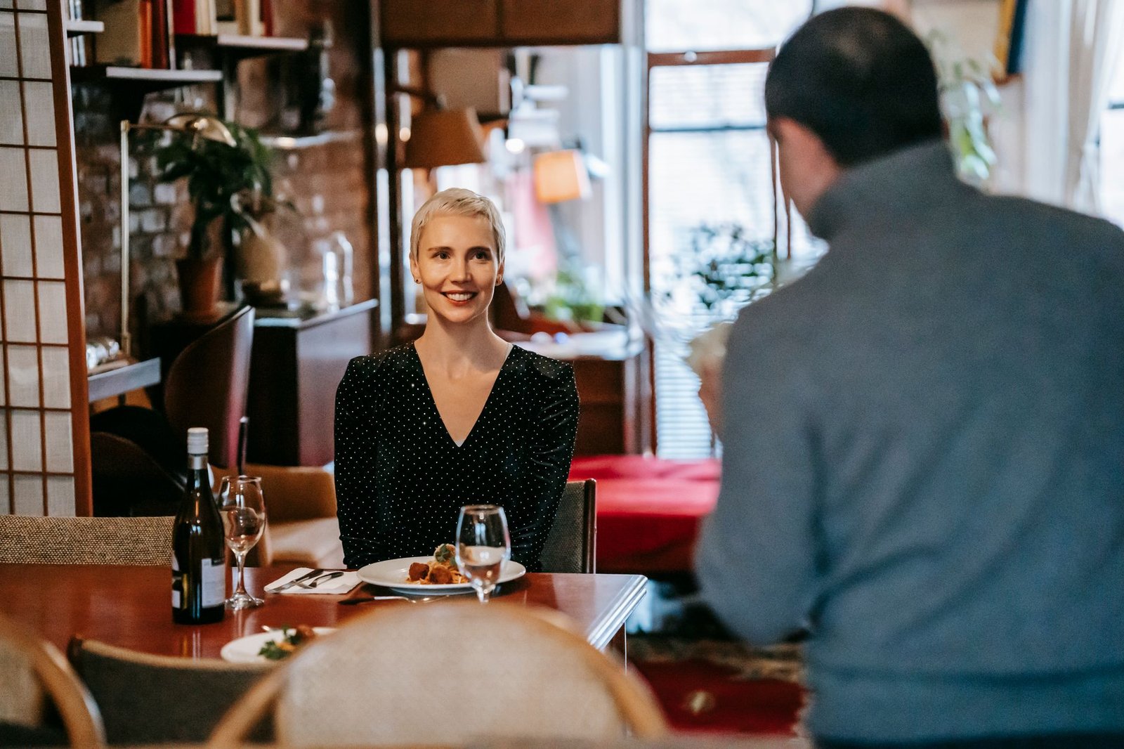 Positive adult couple having date in bright room while man standing with bouquet with flowers near chairs and table with glas